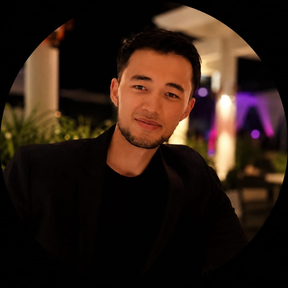 Bek in a black jacket over a black tee, warm indoor evening light on a rooftop lounge behind him