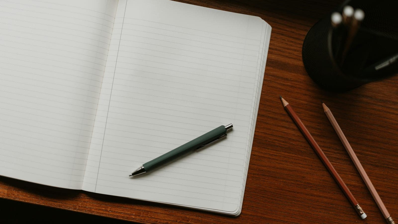An open blank notebook on a wooden desk, a pen and pencils resting on top, ready to be written in